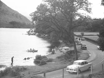 Vista da margem do lago com pessoas de pé, a caminhar, a passear e a andar de barco, a estrada passa à direita da imagem com carros estacionados