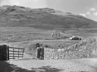 Vista da estrada que passa sobre Hard Knott Pass, duas pessoas de pé perto de um sinal de trânsito e um carro estacionado na berma da relva