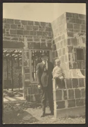 [Homem de pé com criança em casa em construção].