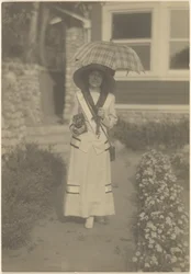 [Mulher em pé no jardim da frente com uma câmara e um guarda-chuva xadrez]
