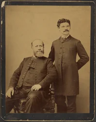 [Homem barbudo não identificado sentado com homem não identificado de bigode em pé]