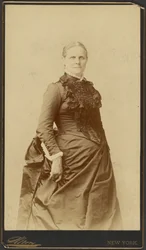 [Mulher não identificada, de pé, com vestido comprido]