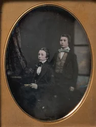 Dois rapazes de gravata borboleta, um sentado segurando um livro, outro em pé, década de 1850.