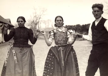 Duas senhoras e um homem, com vestidos tradicionais húngaros, estão de mãos dadas numa dança, segurando algumas velas nas mãos