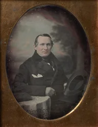 Homem vestindo casaco e luvas, segurando o chapéu, sentado na frente do pano de fundo pintado ao ar livre, década de 1840.