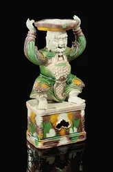Castiçal de espigão com vidrado amarelo ovo e verde espinafre, modelado como um imortal sorridente com barba ondulante e os braços erguidos para sustentar o acessório quadrilobado com espigão, equilibrado sobre a cabeça, Kangxi