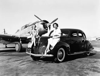 Califórnia: 1939 Duas mulheres sentadas nos pára-lamas de um sedan Lincoln Zephyr de 1939 em um aeroporto com um único avião motor estacionado atrás delas (foto a p/b)
