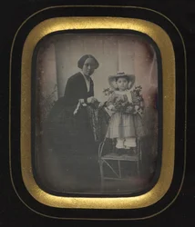 Criança em pé em uma cadeira segurando flores, com a mãe, c. 1855