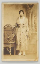 Postal fotográfico de uma mulher ao lado de uma cadeira de madeira, 1904-1918.