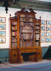 Estante em vidro com grande caixa central decorada com uma moldura interna elaborada incorporando um violino esculpido, ladeada por duas caixas estreitas mais curtas que assentam numa secção inferior de dois armários laterais e uma baía de leitura central.