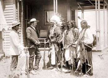 Três Cheyenne vestidos com trajes cerimoniais e armados cumprimentam um branco acompanhado por um intérprete. sd. final do século XIX