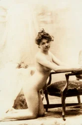 Retrato nu de uma jovem mulher ajoelhada em frente a uma pequena poltrona.