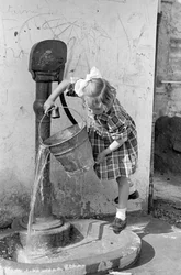 Menina a esvaziar um balde de água numa fonte com bomba, 9 de maio de 1939 (fotografia a preto e branco)