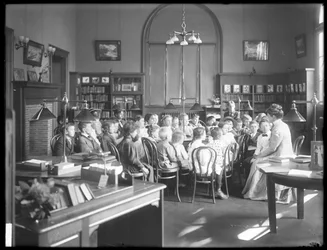 Crianças ouvem uma mulher na sala de leitura de um ramo não identificado da Biblioteca Pública de Queens Borough, c.1910