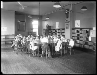 Crianças ouvem uma mulher na sala de leitura de uma filial não identificada da Queens Borough Public Library, c. 1910