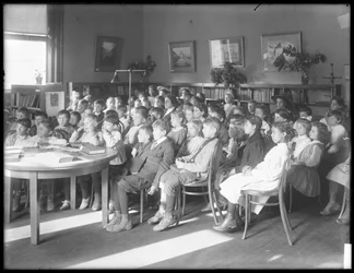 Crianças sentadas, ouvindo na sala de leitura de um ramo não identificado da Biblioteca Pública de Queens Borough, c.1910