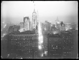 Alto ângulo de visão noturna enevoada da parte baixa de Manhattan olhando para o norte, com o Edifício Singer, o Edifício Woolworth e o Edifício Municipal visíveis, c.1913