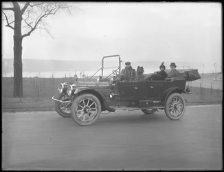 Três pessoas não identificadas e dois cães Chow dirigindo em um carro conversível ao longo de um rio ou lago, c.1912