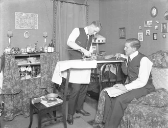 Homem não identificado, demonstrando o uso de um ferro elétrico, assistido por outro homem, c.1913-14