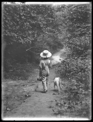 William Gray Hassler de chapéu de palha e macacão, carregando vara de pescar e cesta, andando por um caminho na floresta com um cachorro pequeno (Bessie), 20 de agosto de 1911