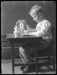 William Gray Hassler sentado em uma pequena mesa comendo uma tigela de cereal de arroz com flocos de Quaker, c.1912