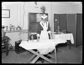 Jovem mulher fantasiada de empregada demonstrando o uso de um ferro elétrico, c.1913-14